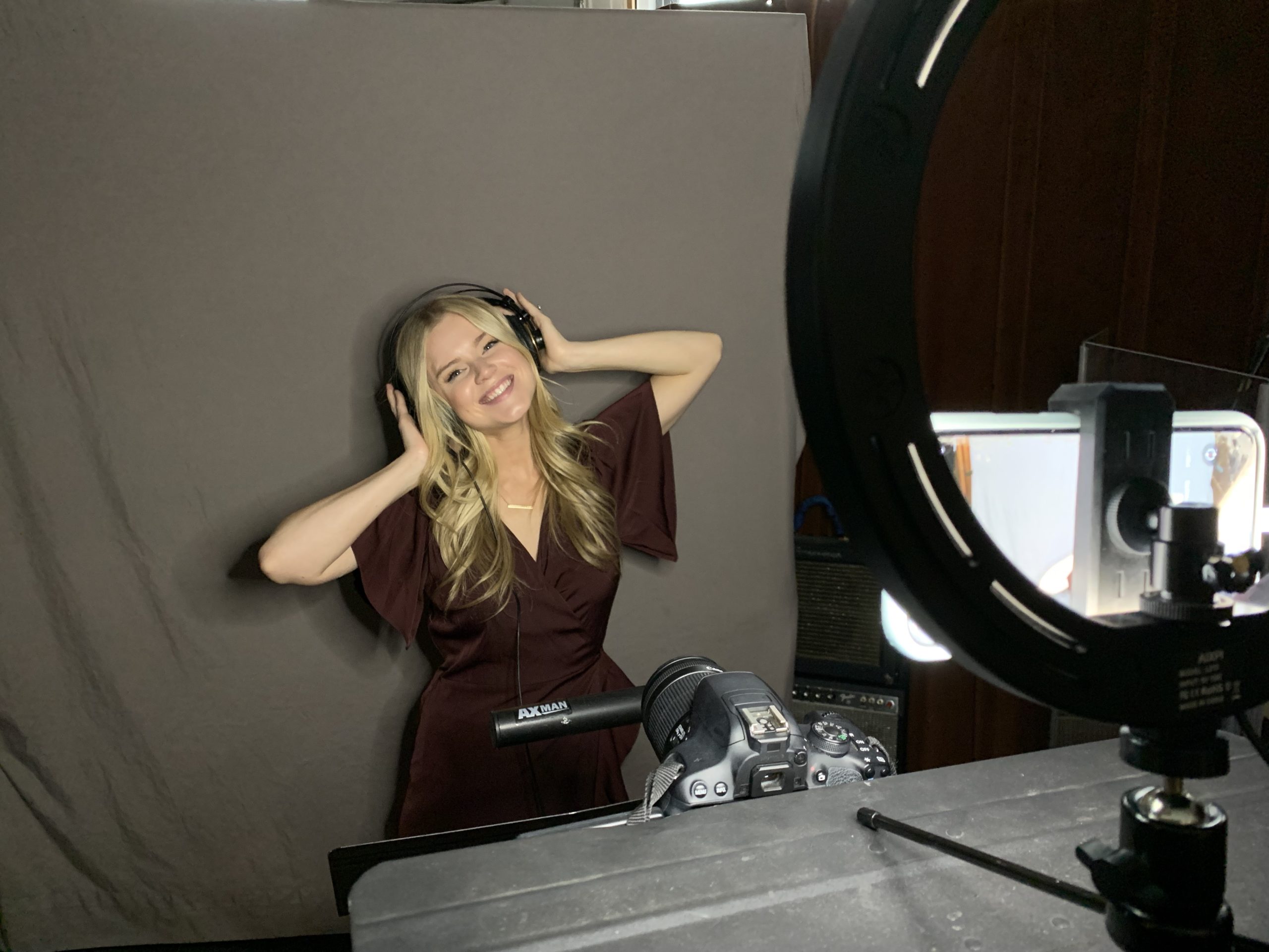 Haley Hilton is a fair-skinned young woman with long wavy blonde hair, wearing a brown short-sleeved dress. She sits in a recording booth with headphones on, in front of a microphone.
