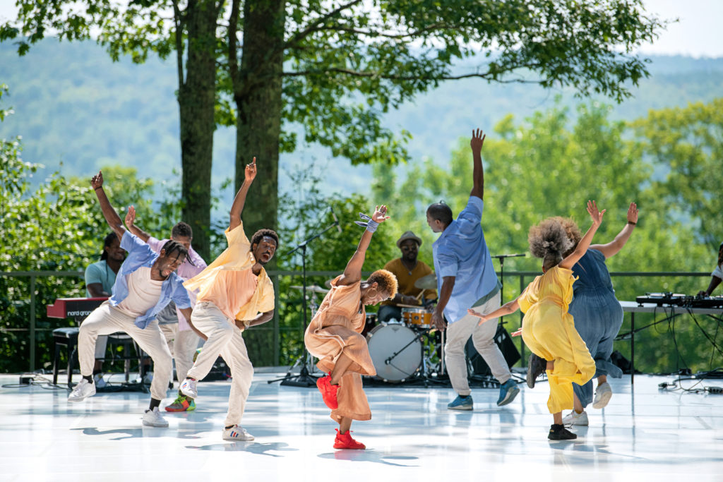 On the outdoor stage at Jacob's Pillow, seven dancers move in a circle, arms outstretched as they lean into their inside legs, the other rising to knee height. LaTasha Barnes is at center, wearing a peach jumpsuit and red-orange sneakers. Musicians sit at a keyboard and drum set at the back corner of the stage.