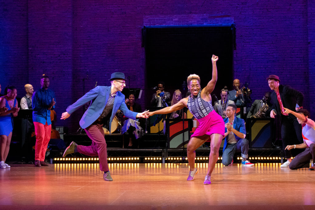 LaTasha Barnes holds the outstretched hand of the dancer beside her, smiling in the direction of the audience as she sits into her opposite hip. Her free arm is raised overhead, as though she's about to snap. Her partner balances on one foot and smiles at her, while musicians and dancers arrayed around them upstage smile and watch.