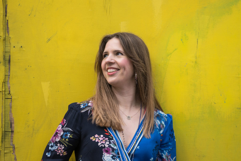 Lucy McCrudden is shown smiling in profile. Her light brown hair is straight and loose around her shoulders. She stands against an unevenly painted yellow wall, wearing a black and blue floral patterned blouse.