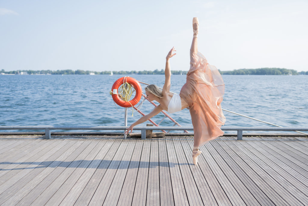 On a pier with sparkling blue water behind it, Genevieve Penn Nabity balances in a six-o'clock penché en pointe. Her blonde hair is loose to her shoulders. Her long peach skirt flutters around her calves.
