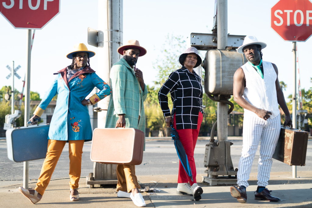 A group of four Black performers pose on a street corner, stop signs on either side. They wear brimmed hats and different combinations of colorfully patterned pieces of suits. Three carry old-fashioned suitcases, while a forth holds an umbrella.