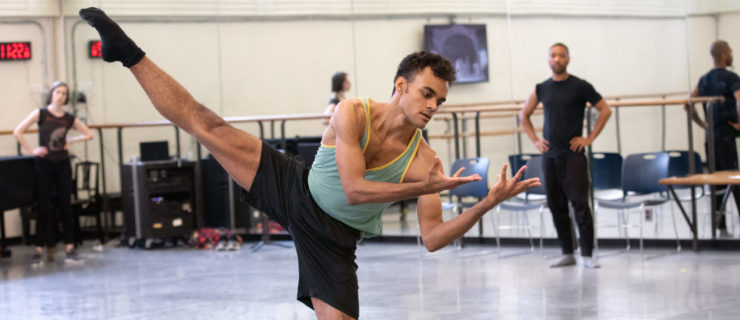 Taylor Stanley balances in plié, his working leg extended above ninety degrees to the side. His torso leans toward his standing leg, slightly contracting as he gazes towards his upturned palms, splayed toward his face, elbows bent towards his chest. He wears a tank top, gym shorts, and socks. Kyle Abraham stands at the front of the rehearsal studio, hands on his hips as he watches.