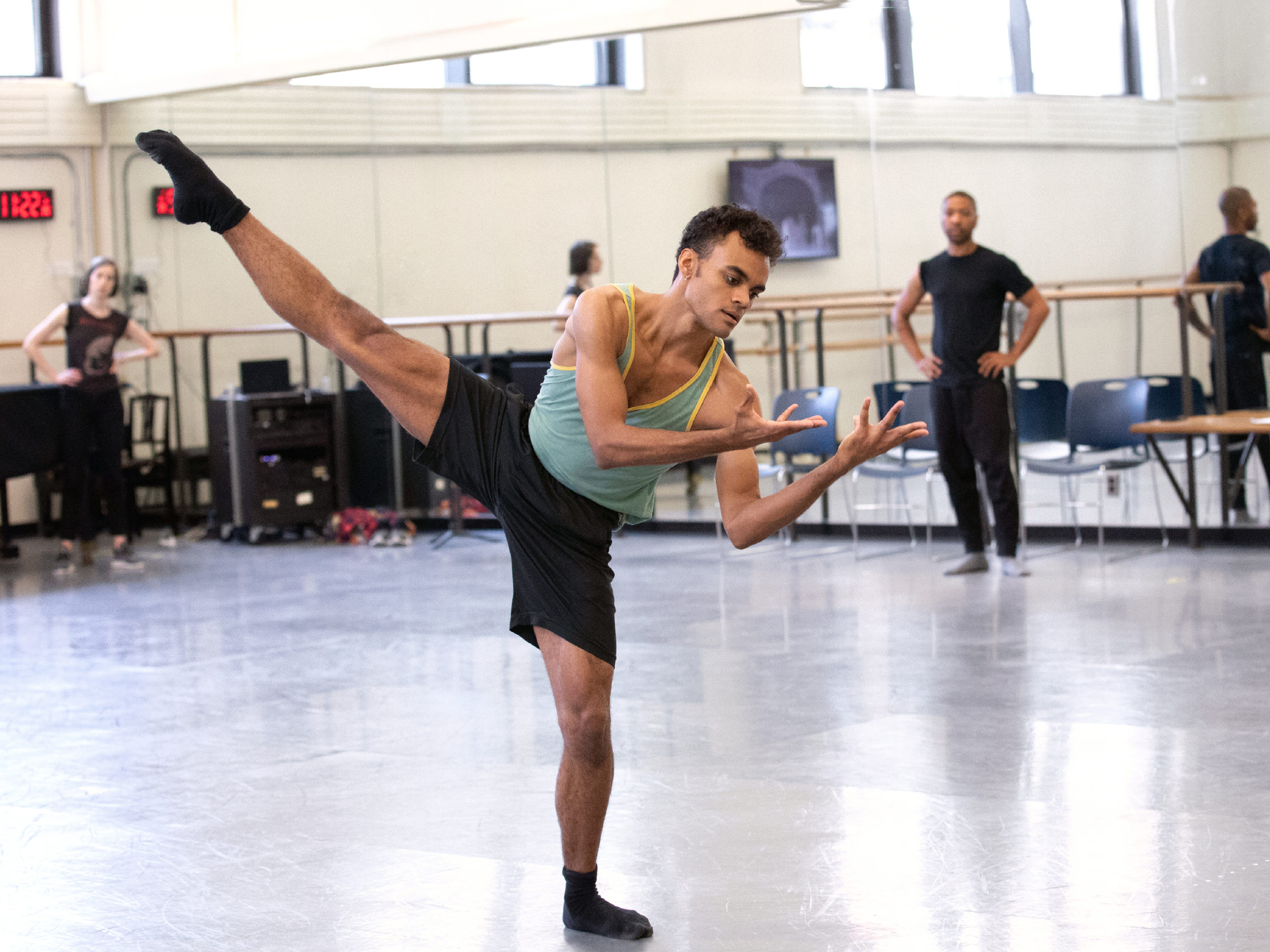 Taylor Stanley balances in plié, his working leg extended above ninety degrees to the side. His torso leans toward his standing leg, slightly contracting as he gazes towards his upturned palms, splayed toward his face, elbows bent towards his chest. He wears a tank top, gym shorts, and socks. Kyle Abraham stands at the front of the rehearsal studio, hands on his hips as he watches.