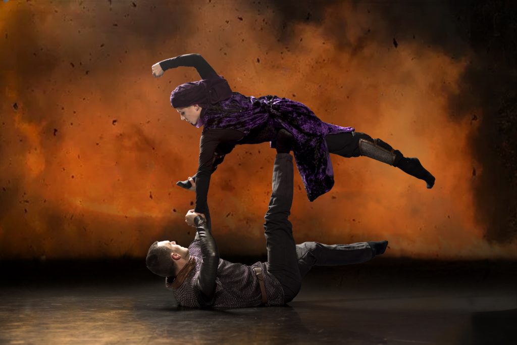 A cloudy orange and black background gives the impression of fire and ash. A fight between two characters. A female dancer in purple dress and headscarf raises a fist as she drifts parallel to the ground, supported by a foot pressed into her hip by the male dancer lying prone beneath her.
