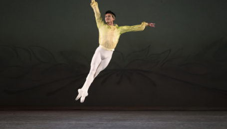 Chun Wai Chan flies through the air, legs in a tight fifth, arms elongated, one beside his ear and the other to the side. He wears white ballet slippers and tights with a pale green, long-sleeved ballet tunic.
