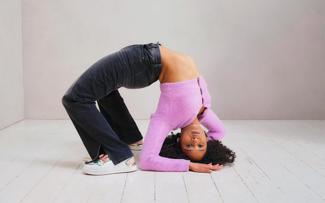 Tania Dimbelolo is a young woman with a medium complexion and curly dark hair. She wears a pink cropped sweater, jeans and white sneakers and rests on her elbows in a backbend.