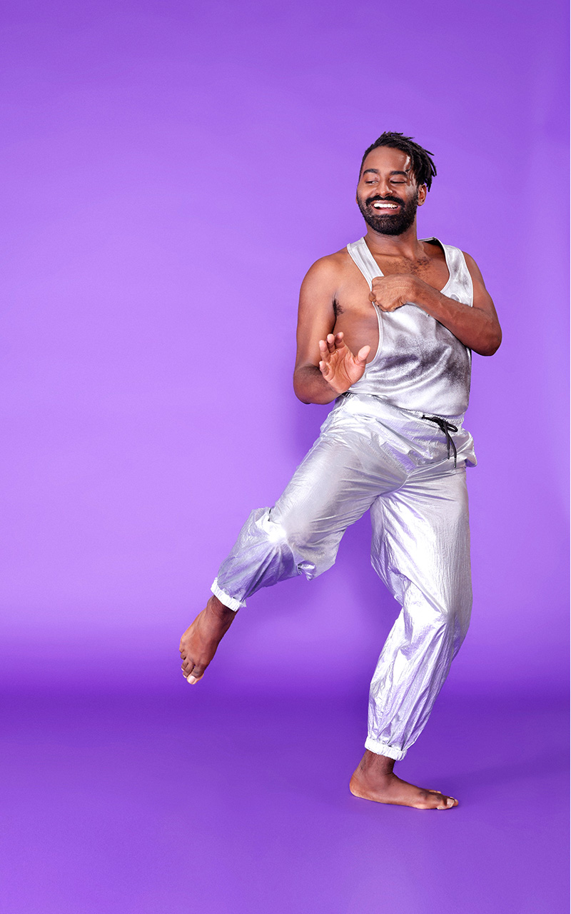 Jerron Herman, a medium-build dark-skinned Black man, here in a silver tank top and pants and bare feet, smiles widely as he looks over his right shoulder and leans over his left leg, with his right leg slightly bent and raised to the side, and his left hand near the strap of his shirt.
