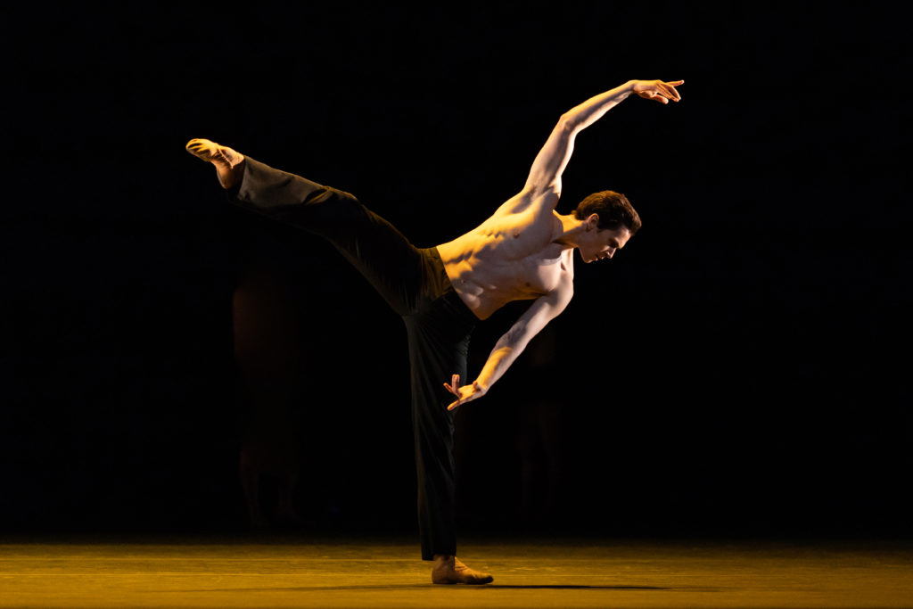 Alone on a shadowy, yellow-lit stage, William Bracewell moves through an off-kilter balance. His right leg is raised in a turned out side attitude as his torso hinges to the left. His gaze is turned downward as his right arm curves in high fifth overhead. He wears black pants and ballet slippers.