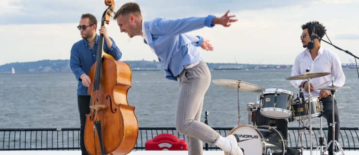 Luke Hickey is caught in profile, arms flying behind him as he taps, grinning, on a shiny tap board. He wears white tap shoes, grey pants, and a sky blue blazer.Behind him on the outdoor stage are a standing bass player and a musician at a drum kit. Beyond the stage, ocean.