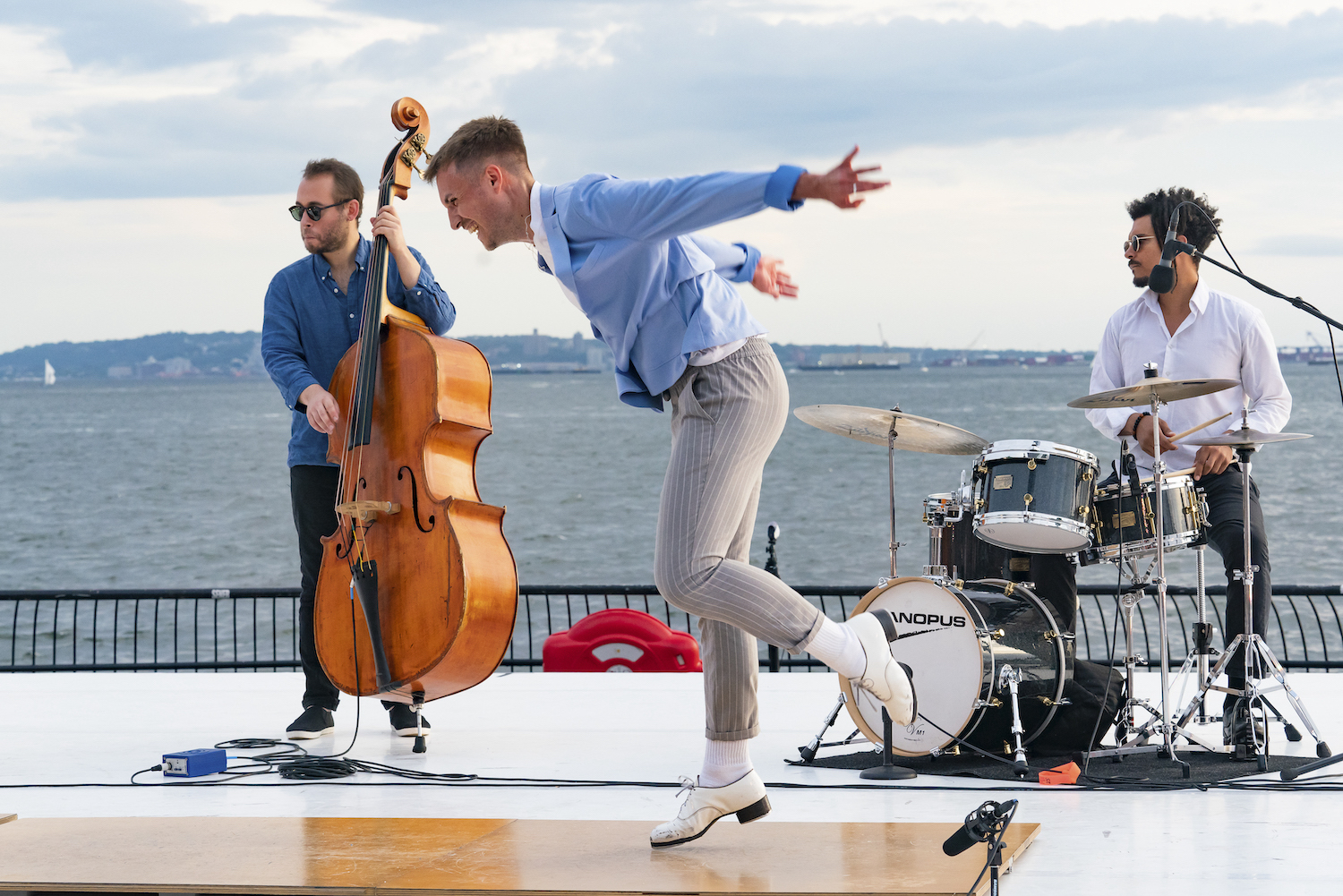 Luke Hickey is caught in profile, arms flying behind him as he taps, grinning, on a shiny tap board. He wears white tap shoes, grey pants, and a sky blue blazer.Behind him on the outdoor stage are a standing bass player and a musician at a drum kit. Beyond the stage, ocean.