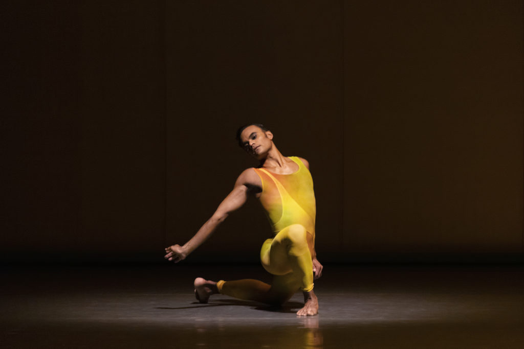 Taylor Stanley is alone onstage, costumed in a unitard in shades of yellow and orange. He lunges, back knee down but toes pressing against the floor. His downstage arm is extended down and back, wrist flexing; he tips his head in that direction, seemingly at ease despite the tension in the gesture.