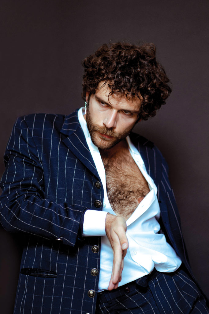 A curly-headed male in a pinstripe suit with the shirt unbuttoned halfway.