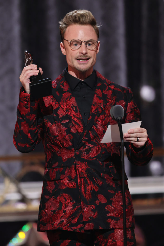 Christopher Wheeldon stands in front of a microphone, holding up a Tony Award statue with one hand and a notecard with his acceptance speech in the other. He wears round glasses and a red and black patterned suit.