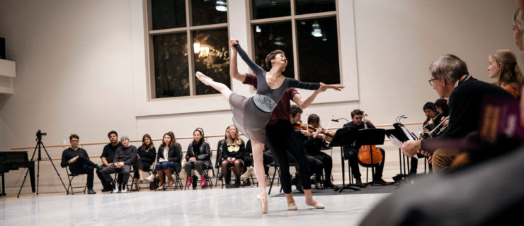 A dancer in pointe shoes, pink tights, a ballet skirt and long-sleeved leotard balances in arabesque, held at the wrists by a partner. Beyond the pair, off the marley, a cluster of musicians. Around the edges of the space, spectators watch from chairs.