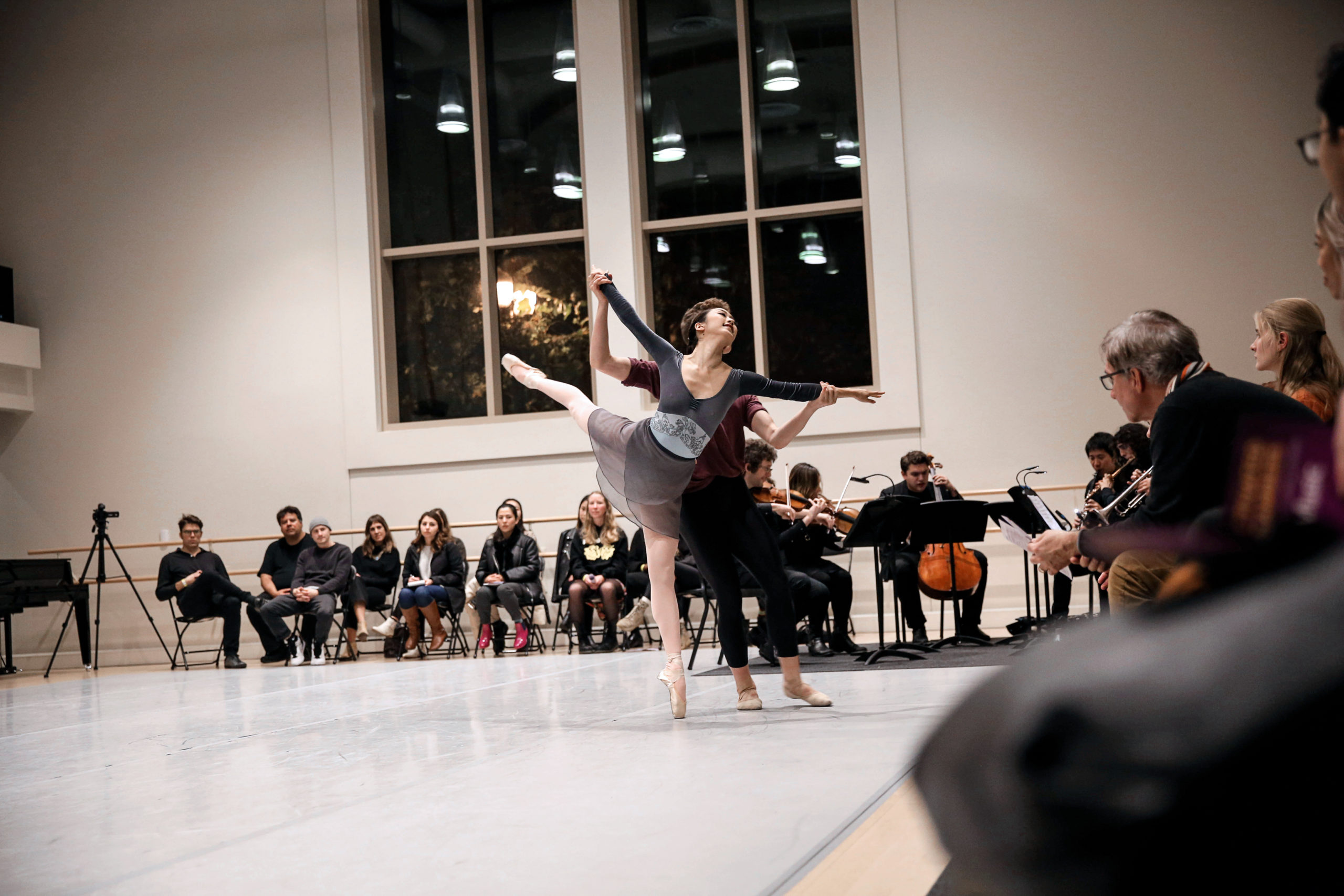 A dancer in pointe shoes, pink tights, a ballet skirt and long-sleeved leotard balances in arabesque, held at the wrists by a partner. Beyond the pair, off the marley, a cluster of musicians. Around the edges of the space, spectators watch from chairs.
