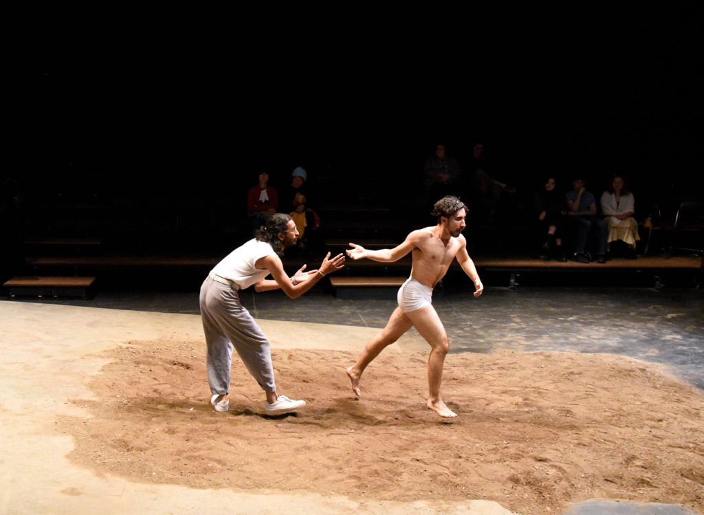 On a pile of dirt, a dancer in white briefs runs away from another who is more fully clothed, having just released his hand. An audience is dimly visible, watching from risers.