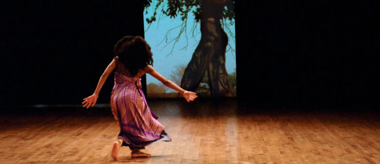 Nosizo Lukhele, a female figure with dark skin and long curly hair in a purple dress with some patterns, is in motion in her back style. On the back screen, a video of herself dancing under a tree is projected.