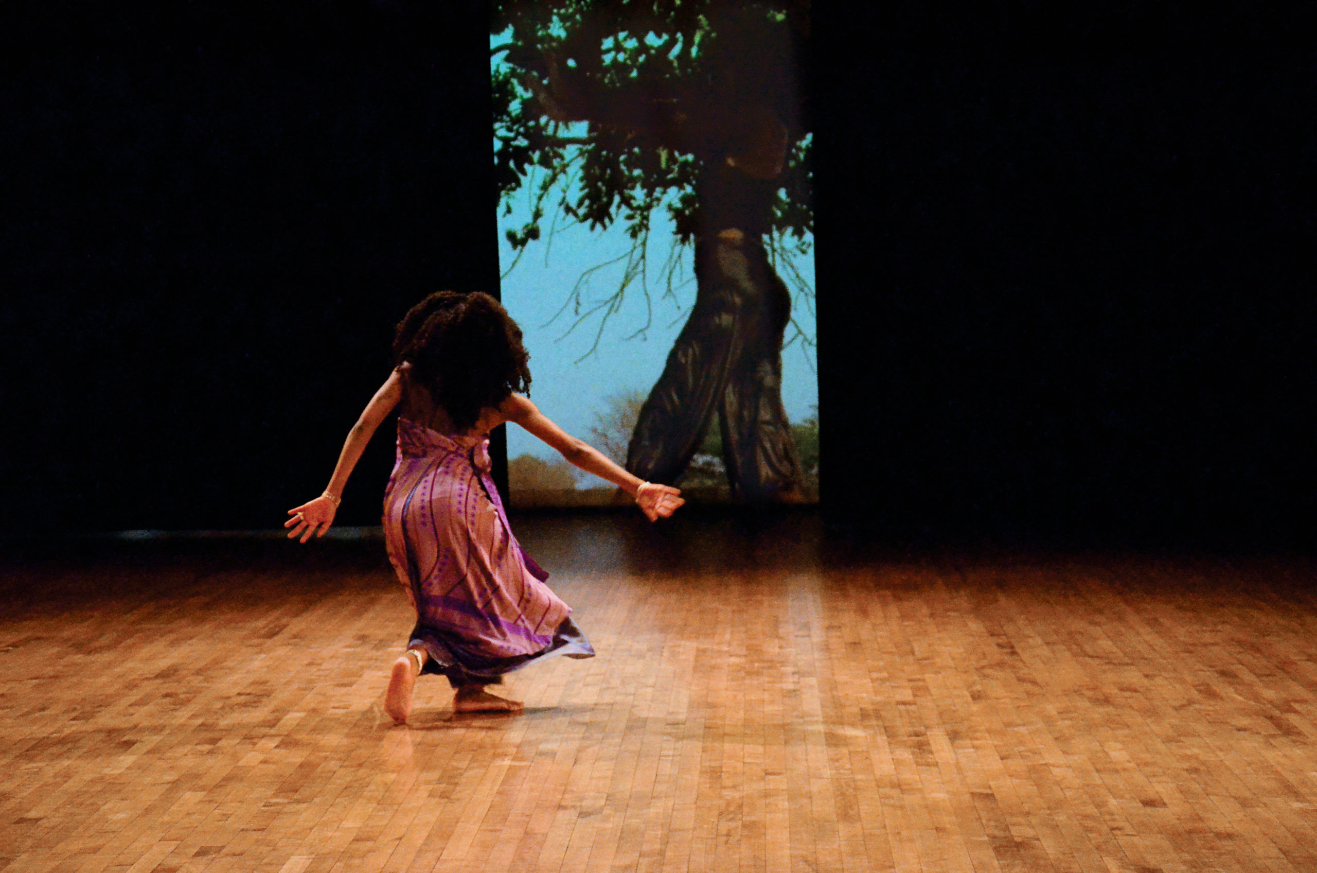 Nosizo Lukhele, a female figure with dark skin and long curly hair in a purple dress with some patterns, is in motion in her back style. On the back screen, a video of herself dancing under a tree is projected.