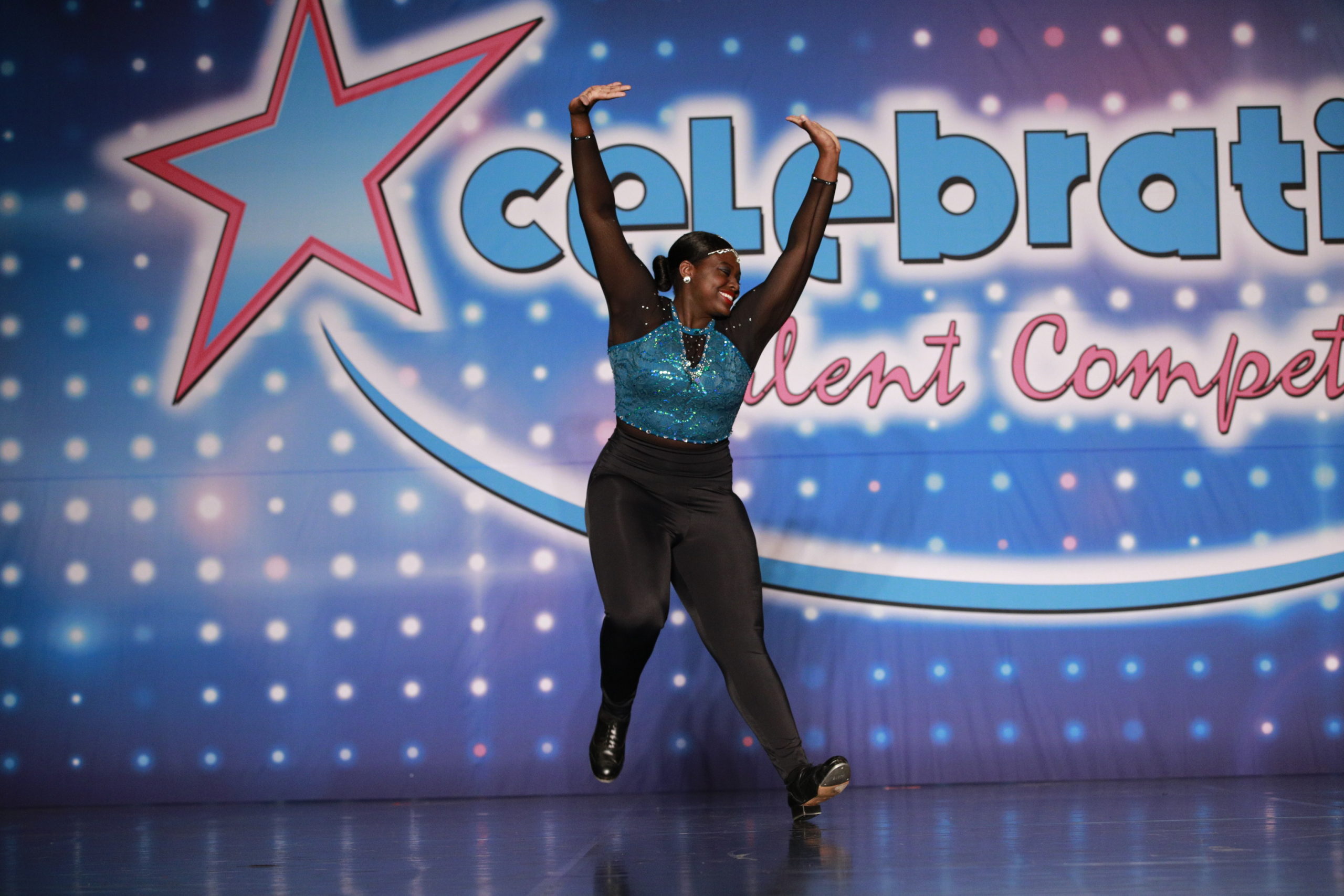 Photo of a young Black female tap dancer, wearing black leggings and a turquoise leotard with long black sleeves, dancing in front of a colorful wall that says "Celebration Talent Competition."