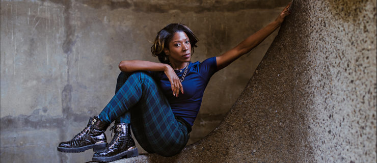 Cira Robinson is a Black woman with ear-length brown hair. She wears a blue short-sleeved t-shirt, blue-black checkered pangs and black shoes, and sits leaning back on a marble wall.