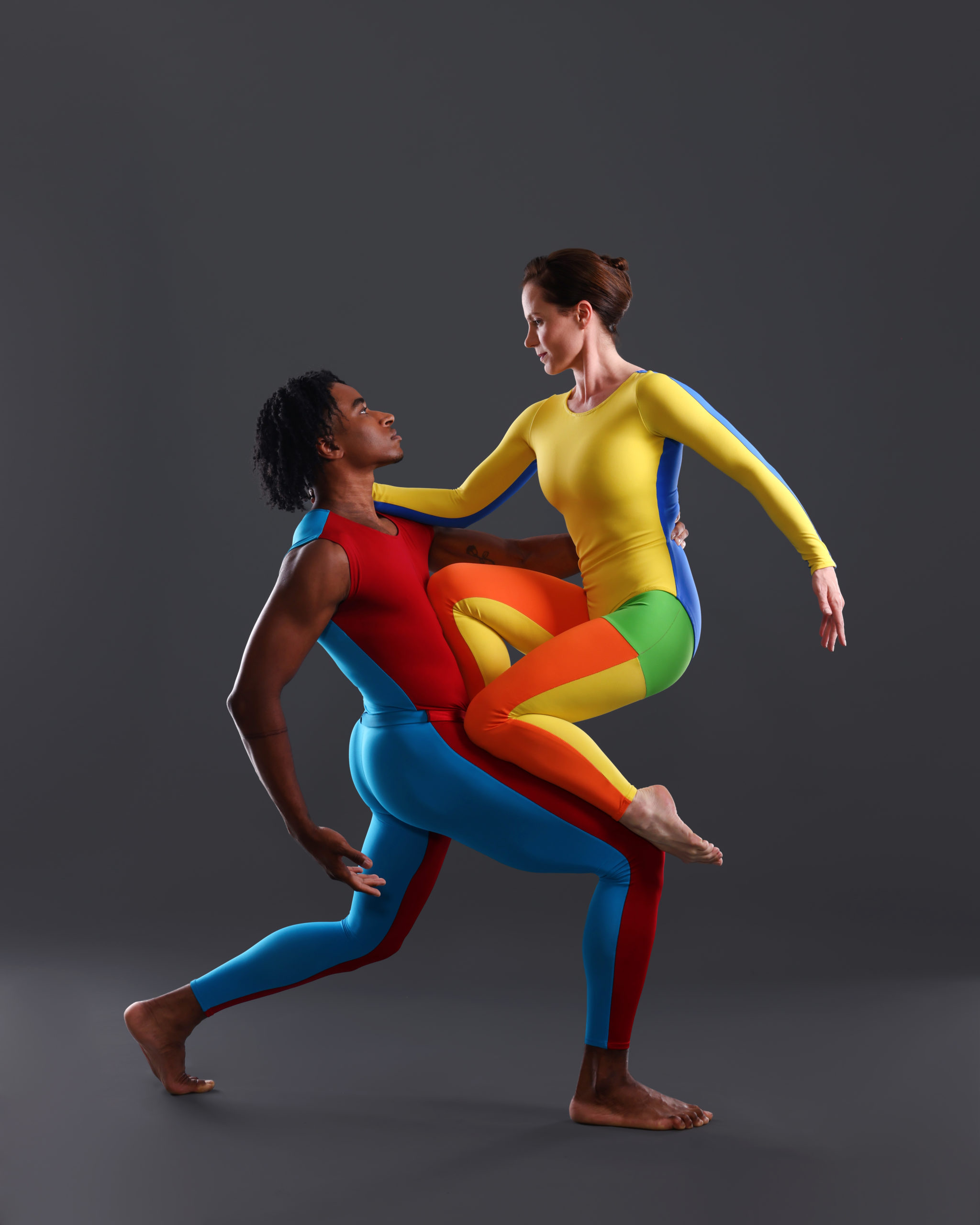 Devon Louis is a black man with chin-length curly hair. Eran Bugge is a fair-skinned woman with long brown hair pulled into a bun. They wear rainbow-colored unitards, and Louis lunges forward on his right leg, supporting Bugge as she kneels on his thigh.