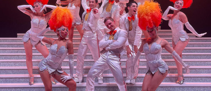 A group of female dancers in sparkling showgirl bodices and orange ostrich-plume headdresses with male dancers in sparkling white tuxedos with orange bow ties.