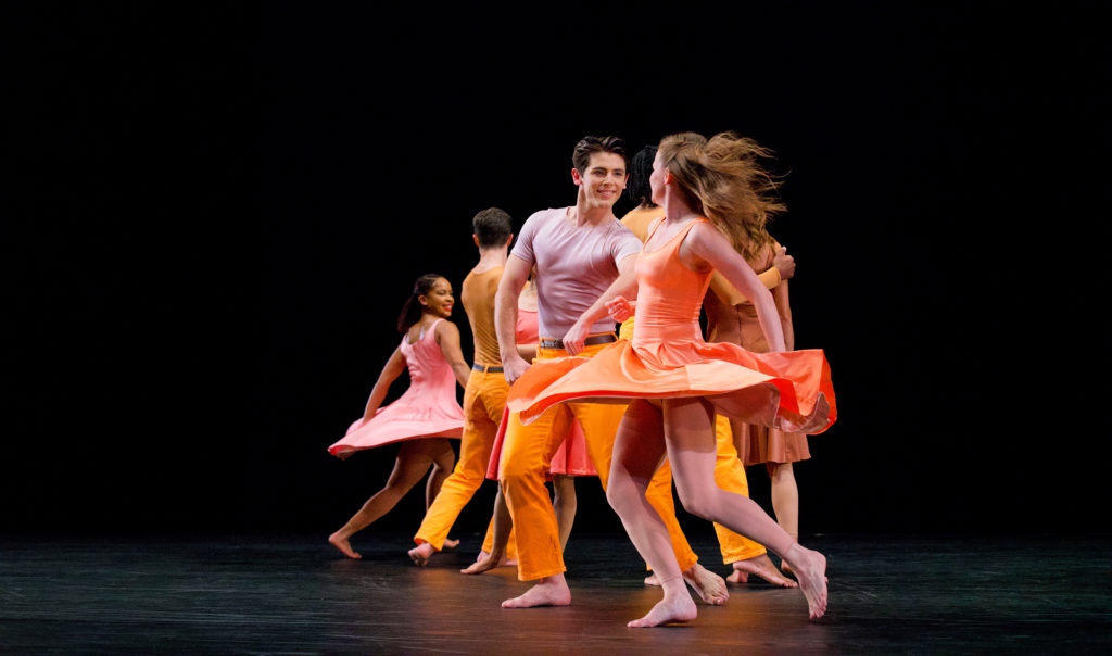 At the front of a line of paired dancers, Jake Vincent offers his arm with a smile to Kristin Draucker, her hair and skirt flying as she spins toward him. The dancers all wear smiles and bright colors reminiscent of a sunrise—sleeveless short dresses for the women, belted pants with well-fitted t-shirts for the men.