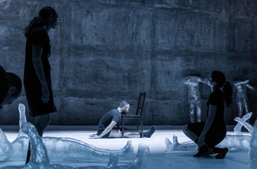 Against a grey, industrial seeming backdrop, a male dancer in t-shirt and shorts sits on the floor with his chin resting on a chair, his legs extended straight beneath it. In the foreground, shadowy dancers stand or kneel amidst translucent models of headless human bodies that lie  or stand around the space.