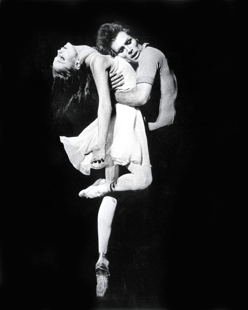 In a black and white archival image, Suzanne Farrell poses en pointe in retiré back, arching back with her arms reaching toward the floor, palms upturned. Her partner's arms are wrapped around her back, his cheek pressed to her upturned chest.