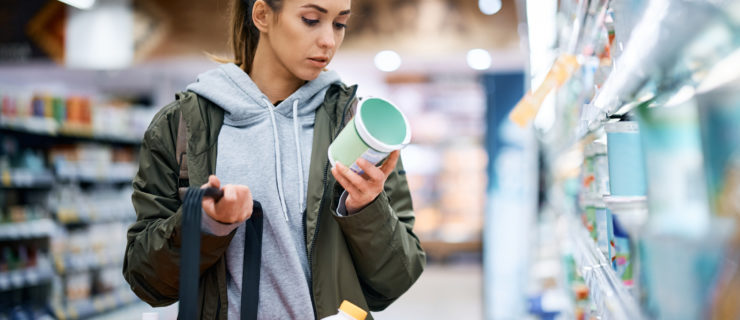 A fare skinned young woman holding a grocery basket and wearing a grey hoodie with a hunter green jacket reads a nutrition label while buying a product in the supermarket.