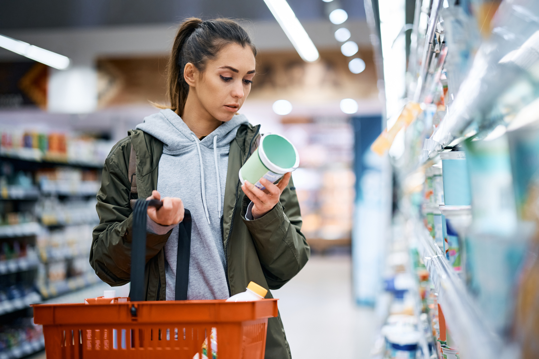 A fare skinned young woman holding a grocery basket and wearing a grey hoodie with a hunter green jacket reads a nutrition label while buying a product in the supermarket.