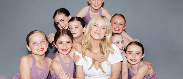 HALEY HILTON is a young fair-skinned blonde woman in a white t-shirt and jeans. She sits cross-legged, surrounded by 8 girls in lavender-colored leotards and skirts.