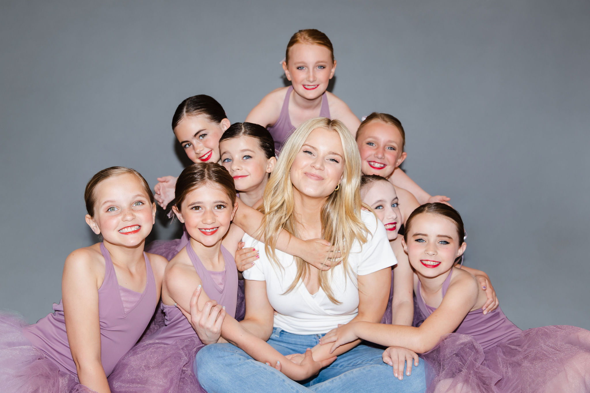 HALEY HILTON is a young fair-skinned blonde woman in a white t-shirt and jeans. She sits cross-legged, surrounded by 8 girls in lavender-colored leotards and skirts.