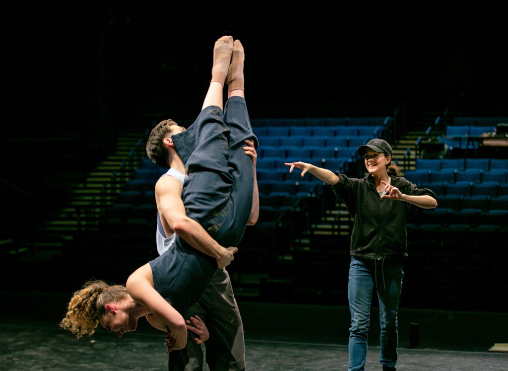 Lauren Lovette smiles as she gestures excitedly towards the pair of dancers she is working with, an arm extended toward them. One dancer is almost upside-down, clutching the thigh of her partner as he wraps his arms around her legs, extended toward the ceiling. All wear rehearsal clothes. In the background, an empty auditorium.