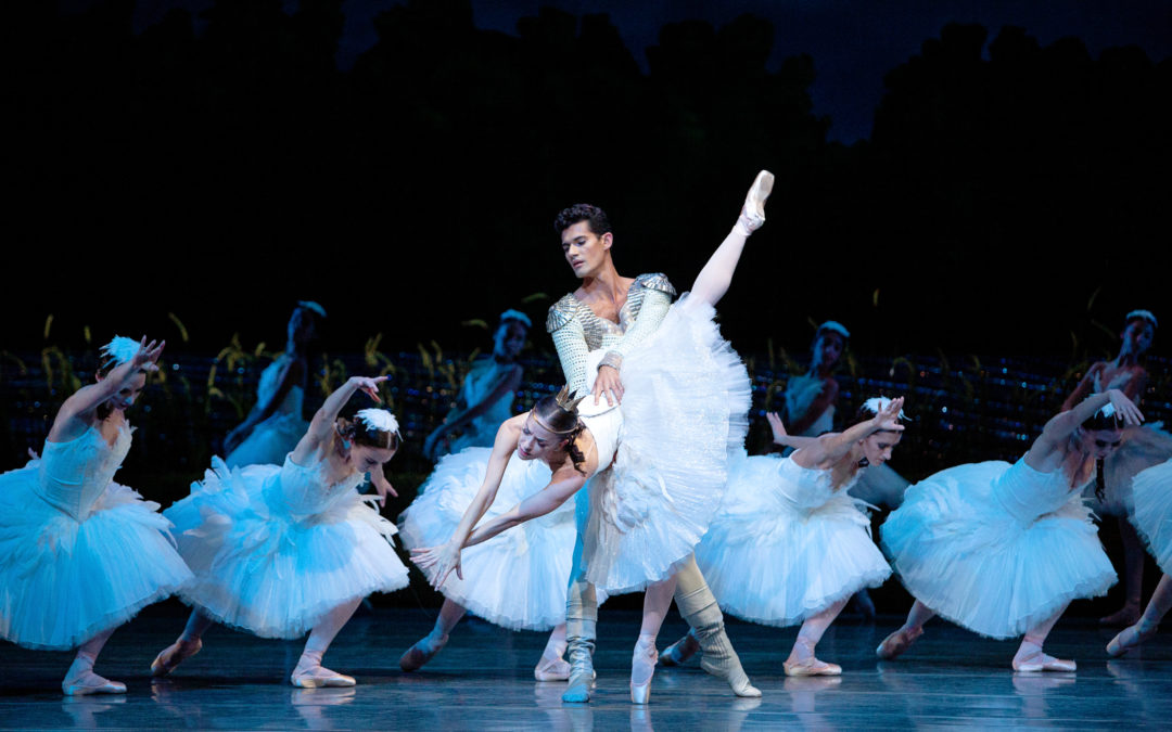Two ballet dancers perform as the White Swan and Prince Siegfried in Miami City Ballet's Swan Lake. The ballerina wears a gold crown and a white tutu, and her partner wears a gold-and-white jacket and beige boots.