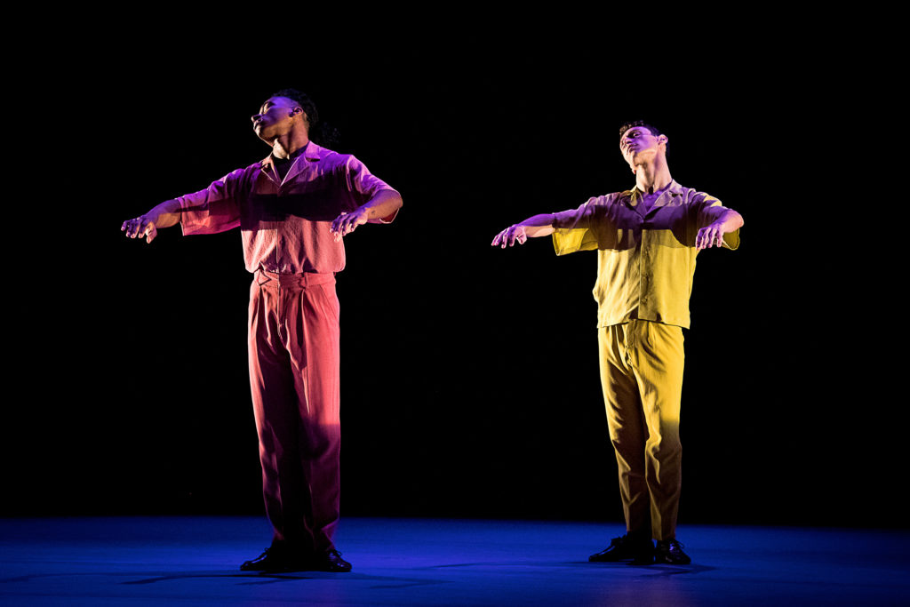 Two male dancers stand onstage in a casual first position, arms raised to rib height and bent in front of them as though resting on a table. Their heads roll back and to their right. One wears head-to-toe pink, the other yellow, shadowed in the stage lighting.