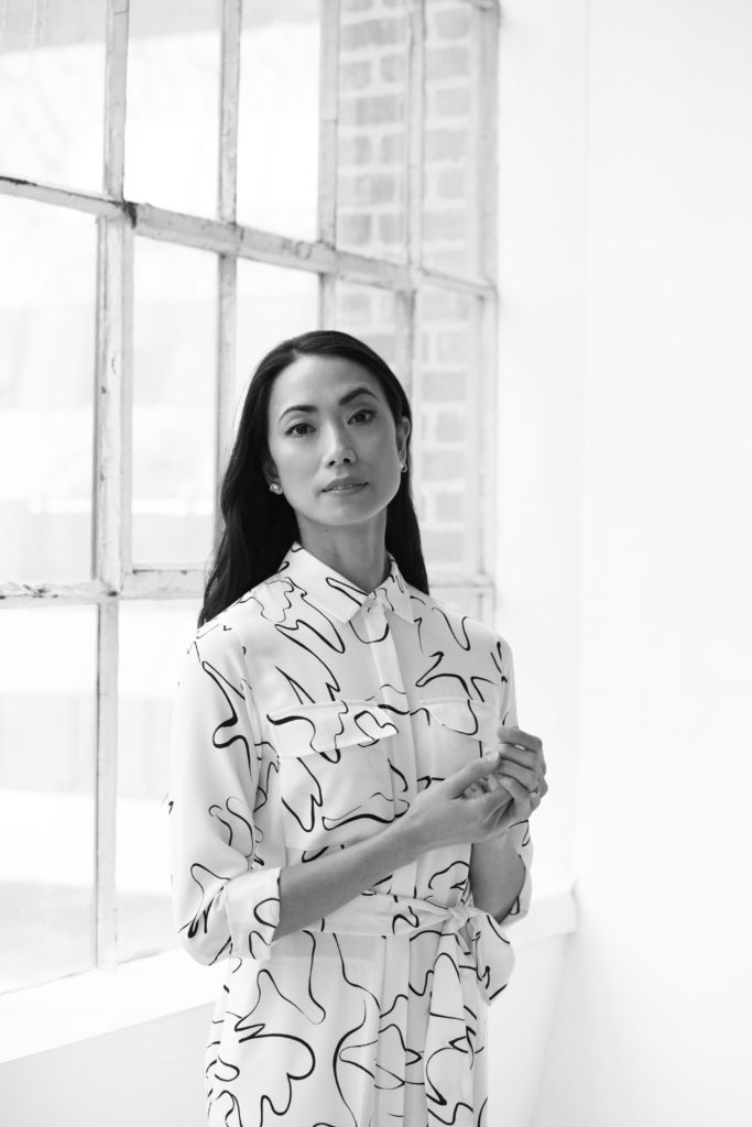 A black and white image of Stella Abrera, who gazes serenely at the camera with one eyebrow raised. She wears a white blouse patterned with abstract black squiggles. Her dark hair is loose behind her shoulders.