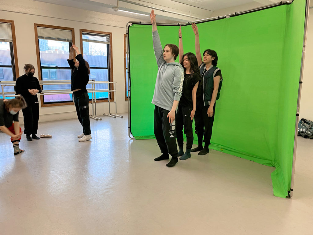 A trio of dancers stand in a line in front of a portable green screen set up in the middle of a studio with their right arms raised overhead. A trio of dancers off to one side separately mark movements or poses.