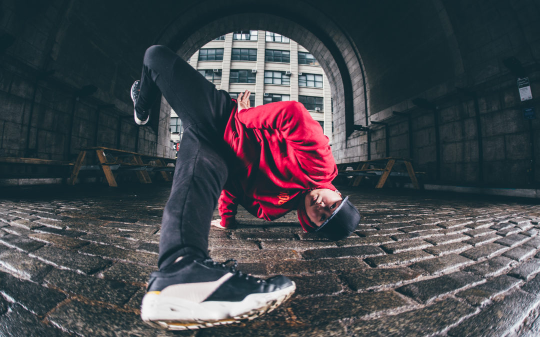 Sunny Choi is an Asian woman wearing a red sweatshirt, black baseball cap and pants, and white sneakers, doing a breaking move on a cobblestone street.