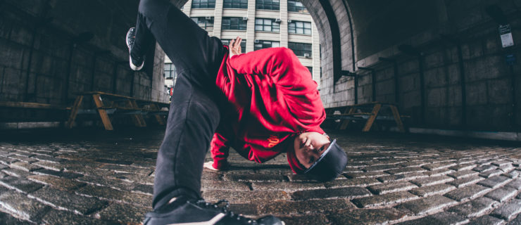 Sunny Choi is an Asian woman wearing a red sweatshirt, black baseball cap and pants, and white sneakers, doing a breaking move on a cobblestone street.