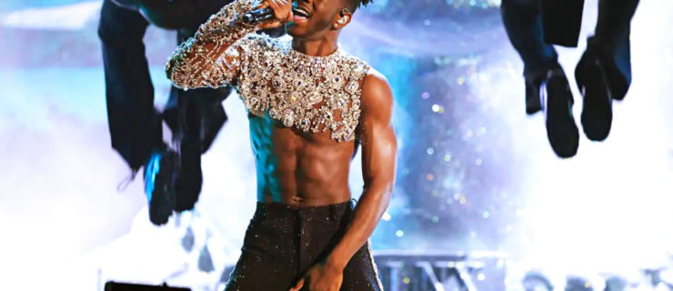 Jaxon Willard is a young Black man with a medium-dark complexion and dark curly hair. He wears a one-sleeve silver-sparkle crop top and black pants and sings into a handheld microphone while two dancers do backflips behind him.