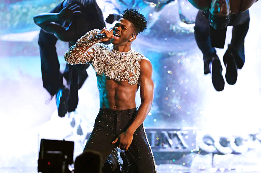 Jaxon Willard is a young Black man with a medium-dark complexion and dark curly hair. He wears a one-sleeve silver-sparkle crop top and black pants and sings into a handheld microphone while two dancers do backflips behind him.