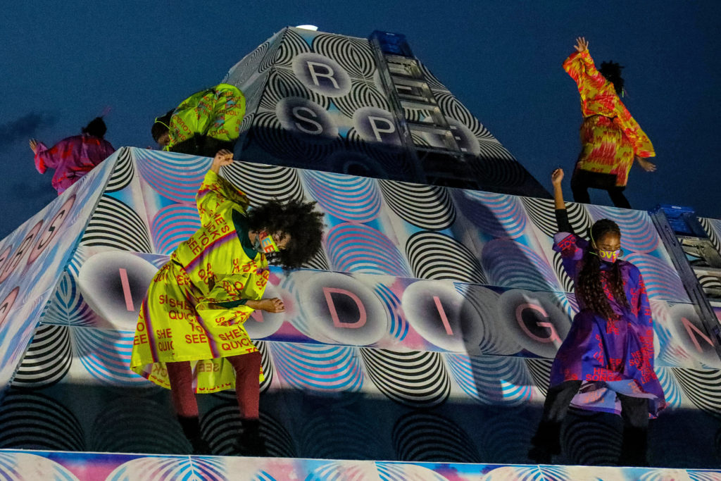 dancers wearing bright costumes dancing on pyramid shaped sculpture 