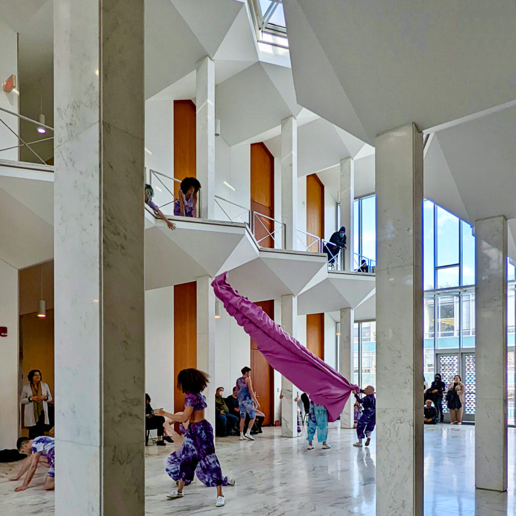 dancers performing in room with marbled floor and pillars, big windows, and white balconies 