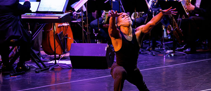 dancer lunging and reaching forward, orchestra playing behind him