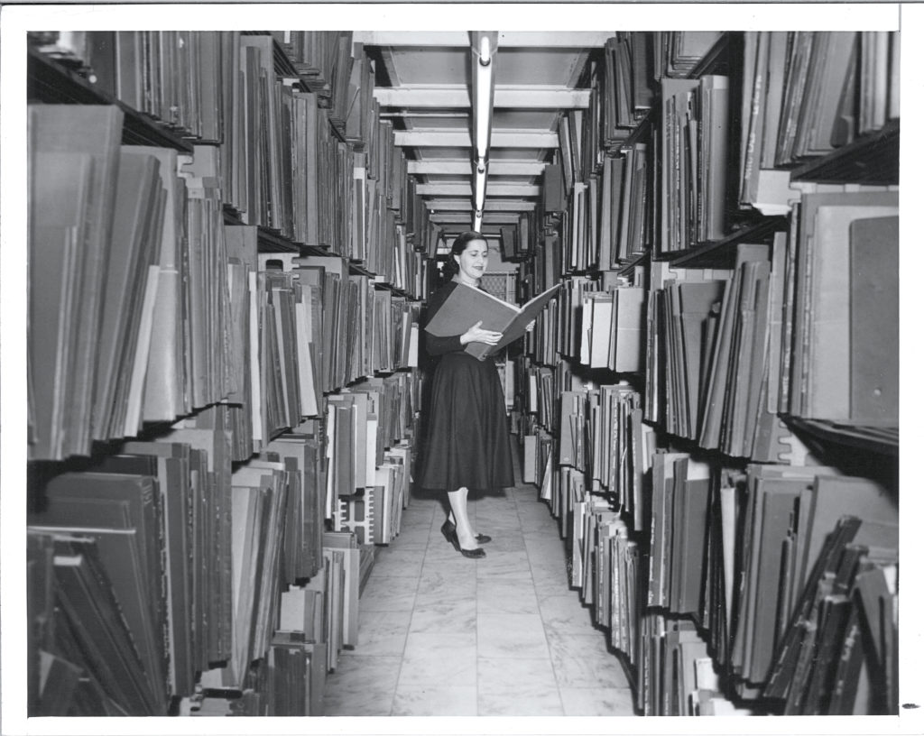 In a black and white archival photo, Genevieve Oswald stands between two packed, towering bookcases. She holds a folio the size of her torso, smiling slightly as she examines its contents. She wears an ankle length dark dress and sensible heels.