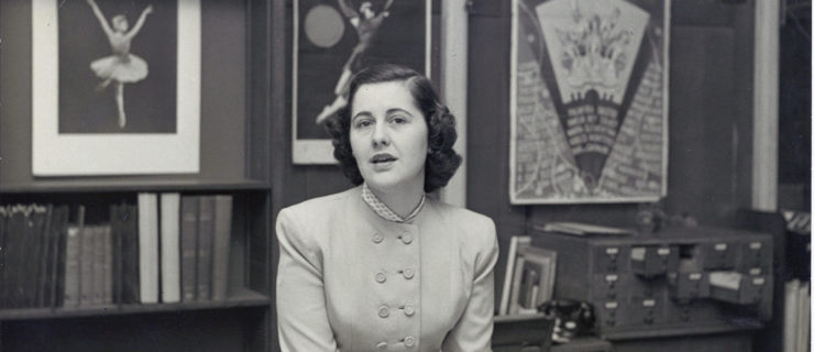 Genevieve Oswald glances toward the camera, mid-speech. Her hands rest on a card catalogue on a full desk; a typewriter is visible to her right. Behind her are shelves with books, and framed posters and portraits of a ballerina, La Meri, and the like. She wears a double breasted, long-sleeved blouse.