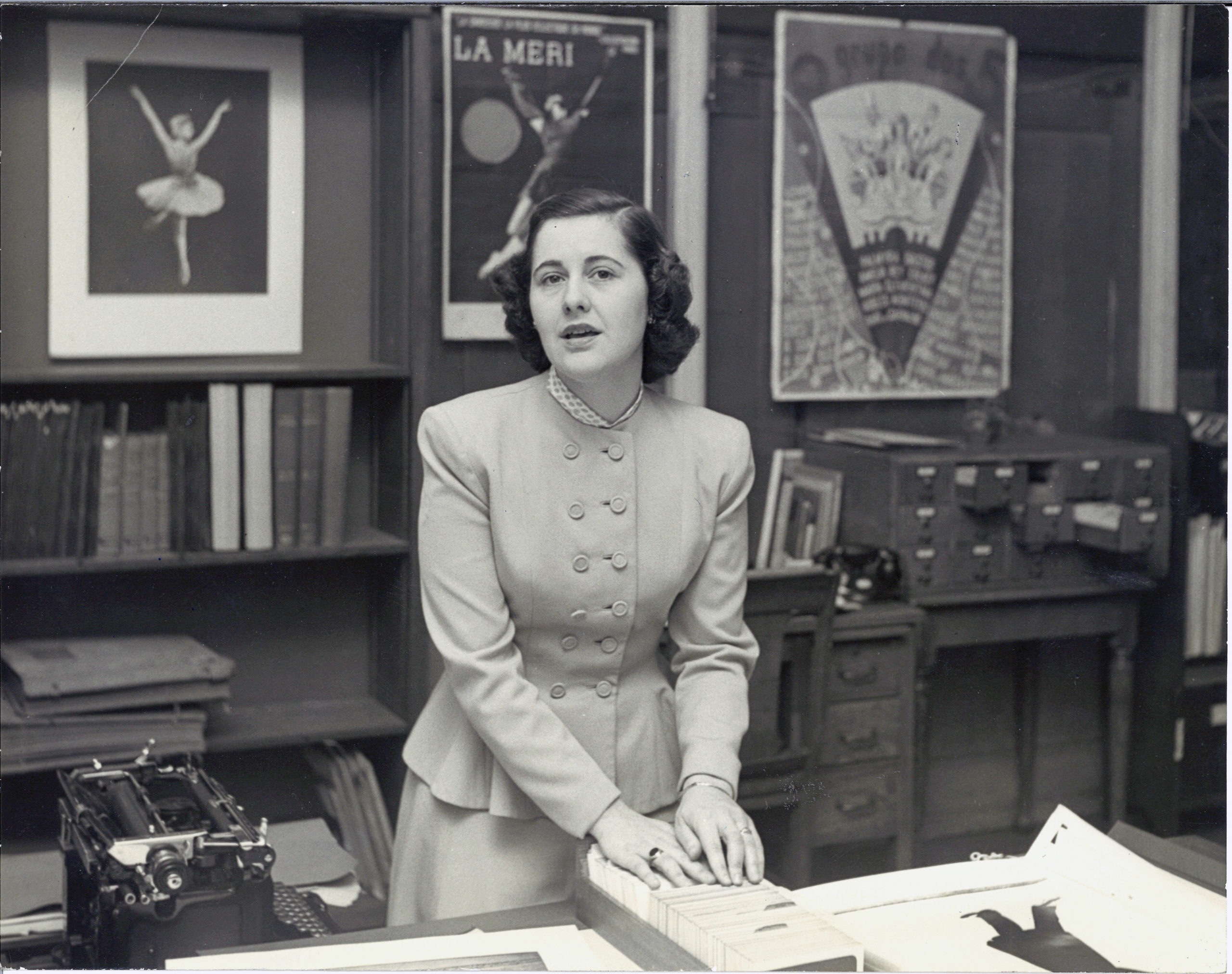 Genevieve Oswald glances toward the camera, mid-speech. Her hands rest on a card catalogue on a full desk; a typewriter is visible to her right. Behind her are shelves with books, and framed posters and portraits of a ballerina, La Meri, and the like. She wears a double breasted, long-sleeved blouse.