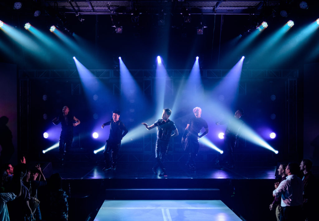 A group of five performers poses together as they sing and dance, the lighting and staging evoking a boy band in performance. Audience members are visible, standing close to the stage.